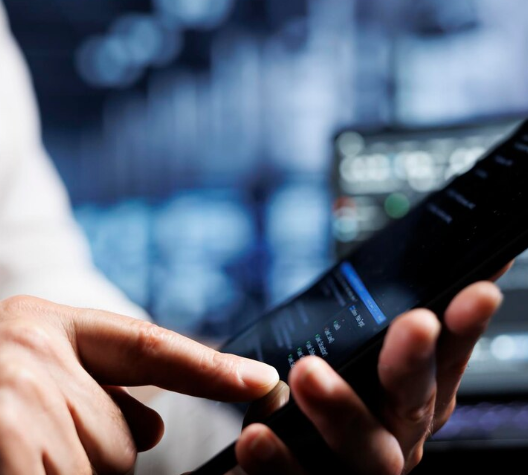 A person's hands holding a smartphone with a dark screen and a blue progress bar at the top. The person's finger is touching the screen. In the background, there is a blurry image of a computer screen with graphs and charts.