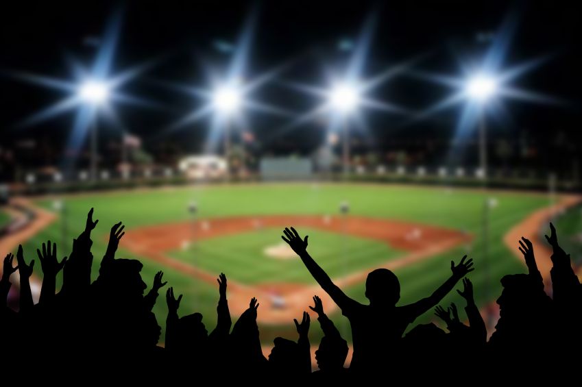 Silhouetted crowd cheering with arms raised in celebration at a baseball game under bright stadium lights.